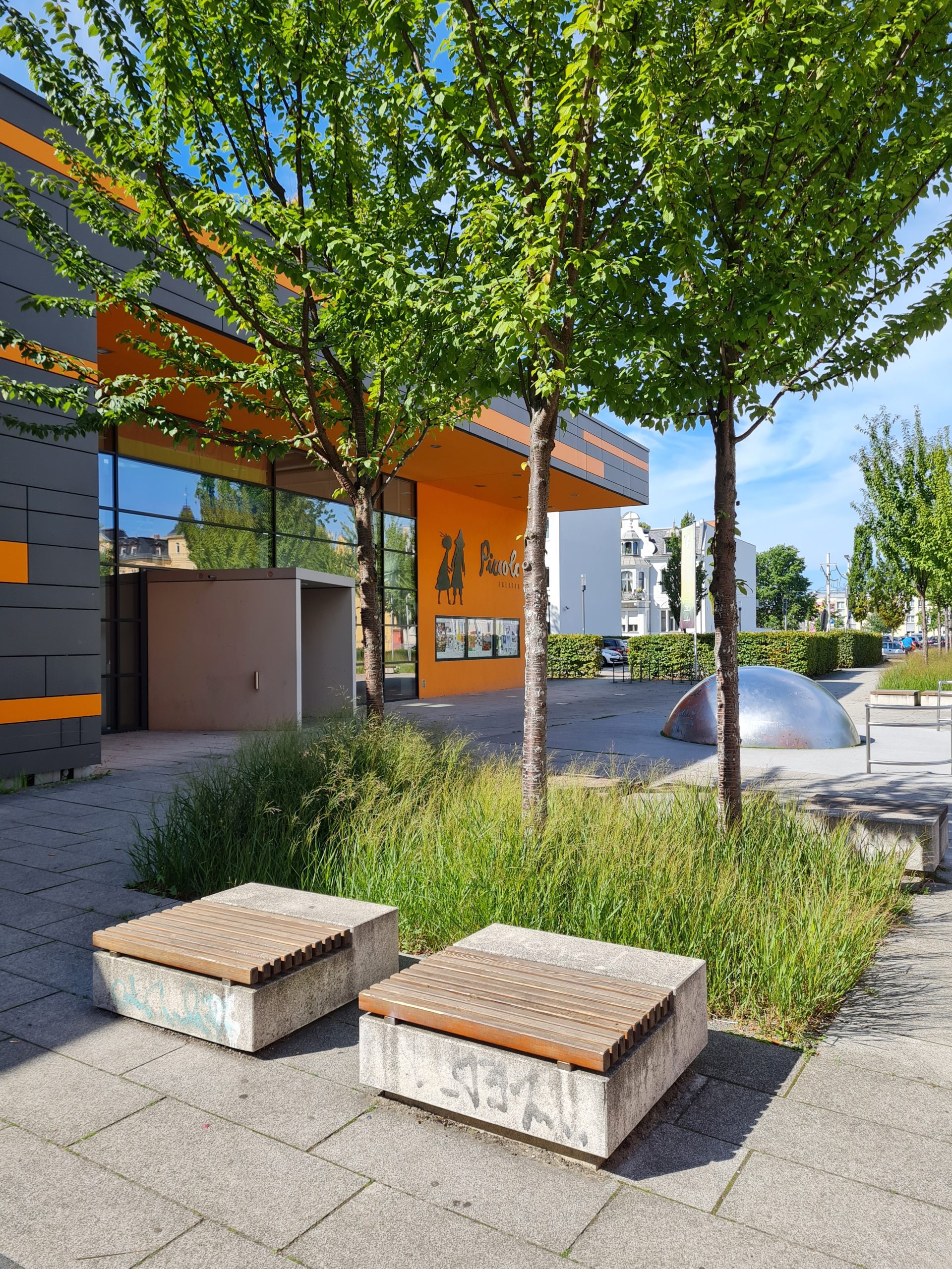 Das Bild zeigt drei Laubbäume auf einem großen Platz vor einer Veranstaltungshalle. Unter den Bäumen befindet sich eine Baumscheibe, die mit Gräsern bepfanzt ist. Davor stehen zwei Sitzmöglichkeiten aus Beton mit Holzverkleidung.